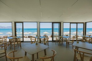 A beachside restaurant with tables and chairs, large windows, and beach umbrellas.