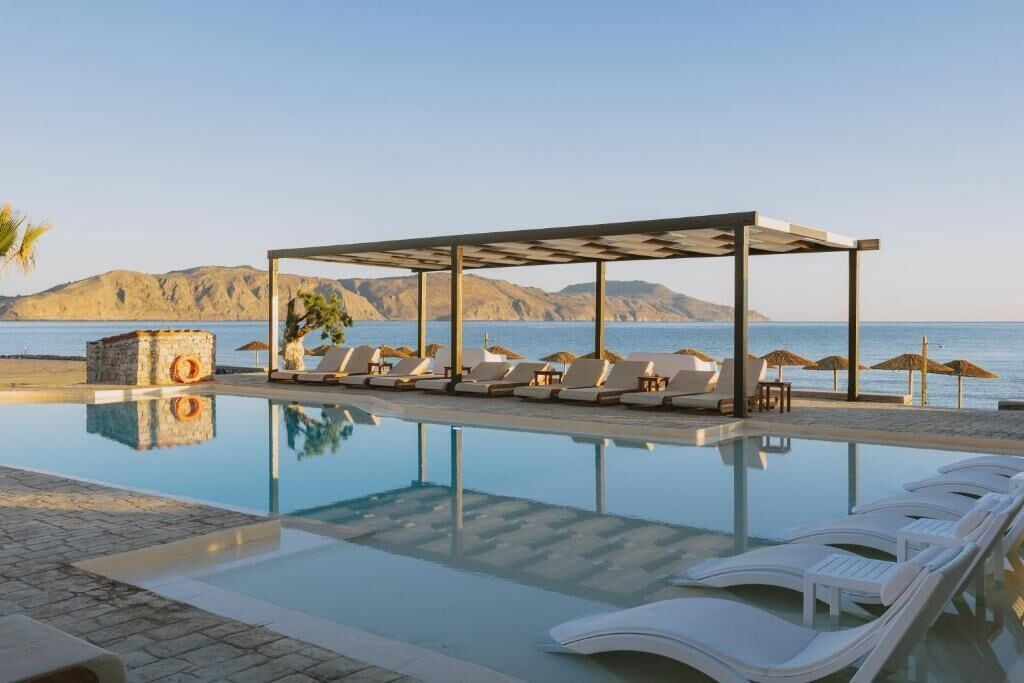 A poolside area with lounge chairs and umbrellas, a stone structure, and a view of the sea and mountains.