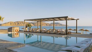 Un espace aménagé au bord de la piscine, avec des chaises longues et des parasols, une structure en pierre et une vue sur la mer et les montagnes.