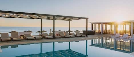Uma área de piscina com espreguiçadeiras e guarda-sóis, um espaço de estar coberto e vista para o mar.