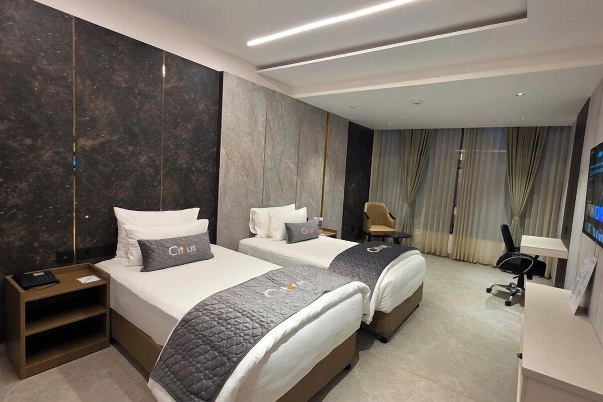 A hotel room with two beds, a desk, a chair, a TV, and a large window with curtains.