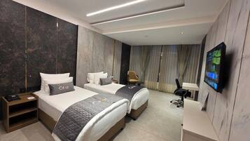 A hotel room with two beds, a desk, a chair, a TV, and a large window with curtains.