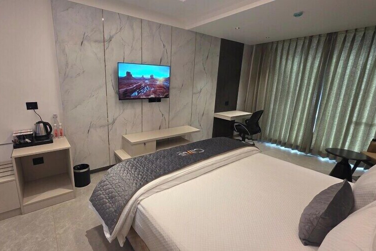 A modern hotel room with a large bed, a TV mounted on the wall, a desk, and a seating area.