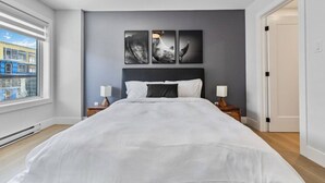 Un dormitorio moderno con una cama grande, dos mesitas de noche, vistas de edificios a través de una ventana y tres cuadros enmarcados en la pared.