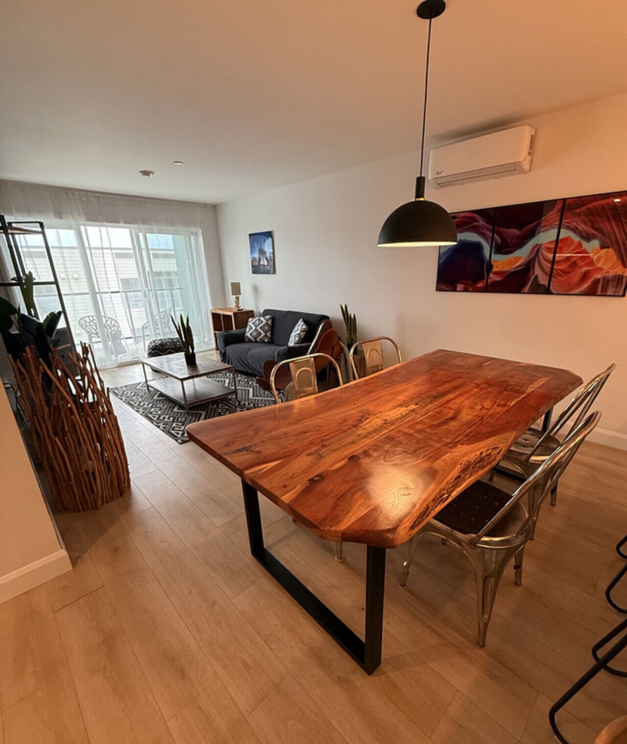 A dining area with a wooden table, metal chairs, a hanging light fixture, and a view of a living room with a sofa and a painting.