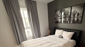 Un dormitorio con una cama, cortinas grises, una mesita de noche, una lámpara y un cuadro de elefantes en la pared.