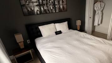 A hotel room with a large bed, a bedside table with a lamp, a robe hanging on the wall, and a painting above the bed.