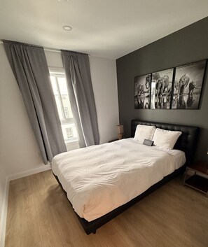 A bedroom with a bed, grey curtains, a bedside table, a lamp, and a wall with elephant artwork. - Prisme (Regina)