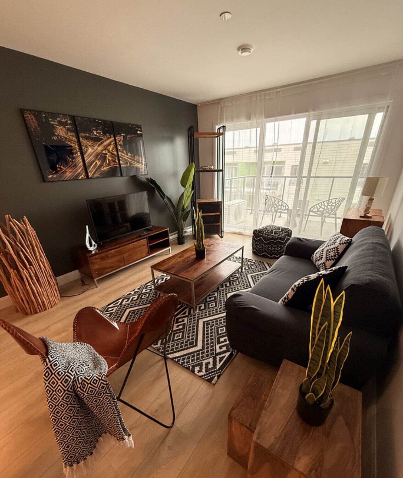A modern living room with a sofa, coffee table, and TV stand. There is a large window with curtains, a plant, and a patterned rug.