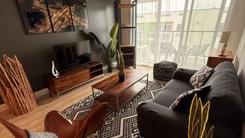 A modern living room with a sofa, coffee table, and TV stand. There is a large window with curtains, a plant, and a patterned rug.
