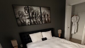 Una habitación de hotel con una cama grande, una mesita de noche con lámpara, una bata colgada en la pared y un cuadro sobre la cama.