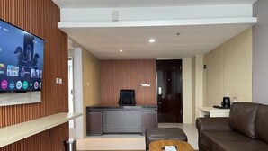 A modern hotel room with a wooden panel wall, a flat-screen TV mounted on it, a reception desk, a brown leather sofa, and a wooden coffee table. - Gulala Azana Hotel and Resort Guci Tegal (Songgom)