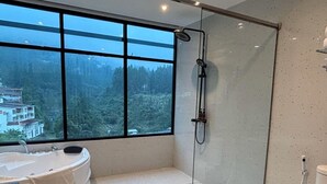 A modern bathroom with a glass-enclosed shower, a freestanding bathtub, and a large window offering a view of a forested area. - Gulala Azana Hotel and Resort Guci Tegal (Songgom)