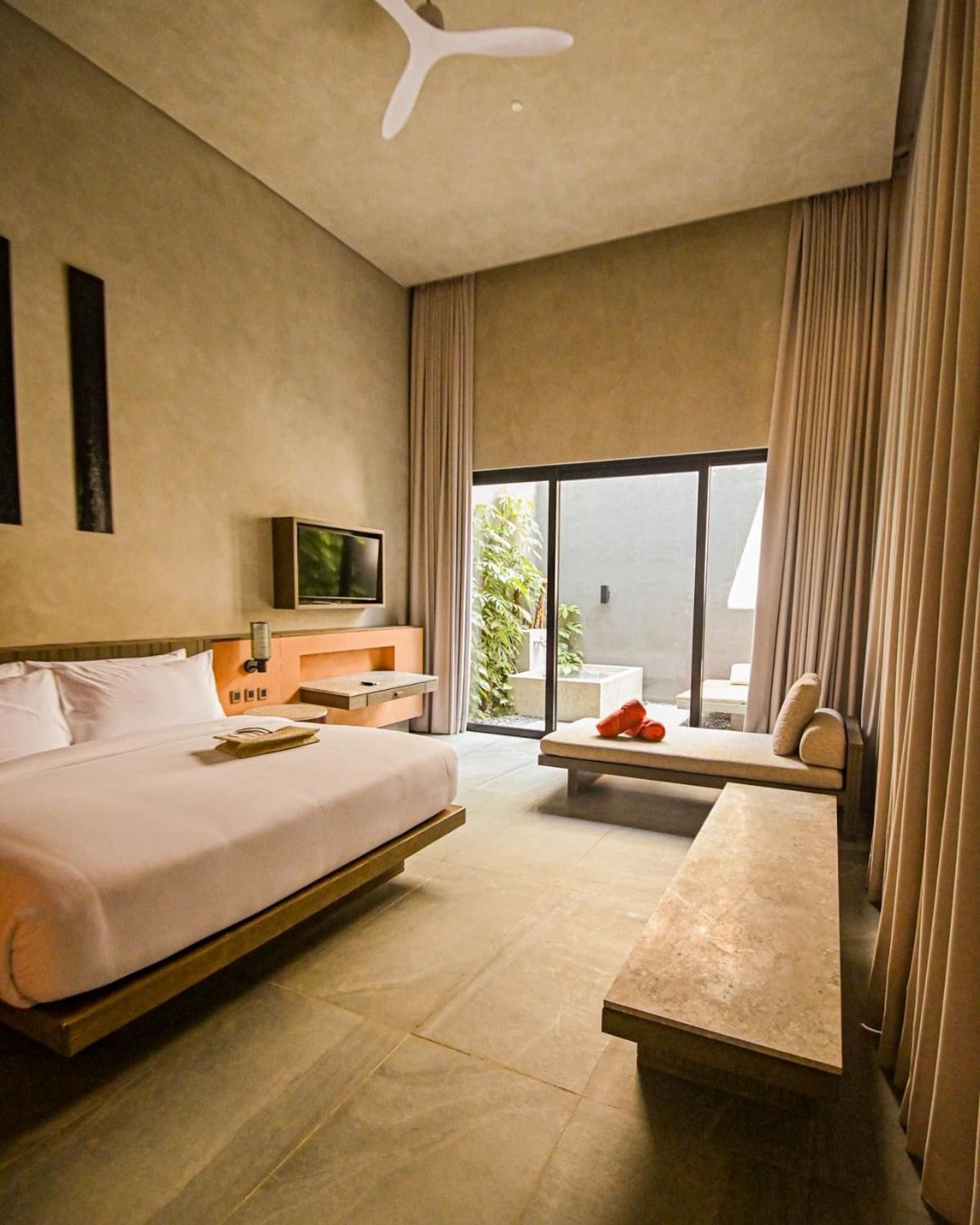 A modern hotel room with a large bed, a flat-screen TV, a desk, and a view of the outdoors.