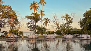 An outdoor dining area with tables and chairs by a body of water, surrounded by palm trees and a clear sky. - Amorita Resort & Spa (Puerto Princesa)