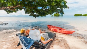 A beach scene with a person lying on a lounge chair under a tree, a red kayak, and a clear view of the water. - Amorita Resort & Spa (Puerto Princesa)