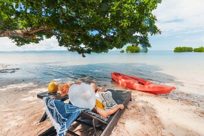 Une scène de plage avec une personne allongée sur une chaise longue à l’ombre d’un arbre, un kayak rouge et une vue dégagée sur l’eau.