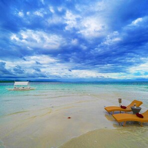 Una playa con agua turquesa cristalina, un bote y dos tumbonas con cojines.