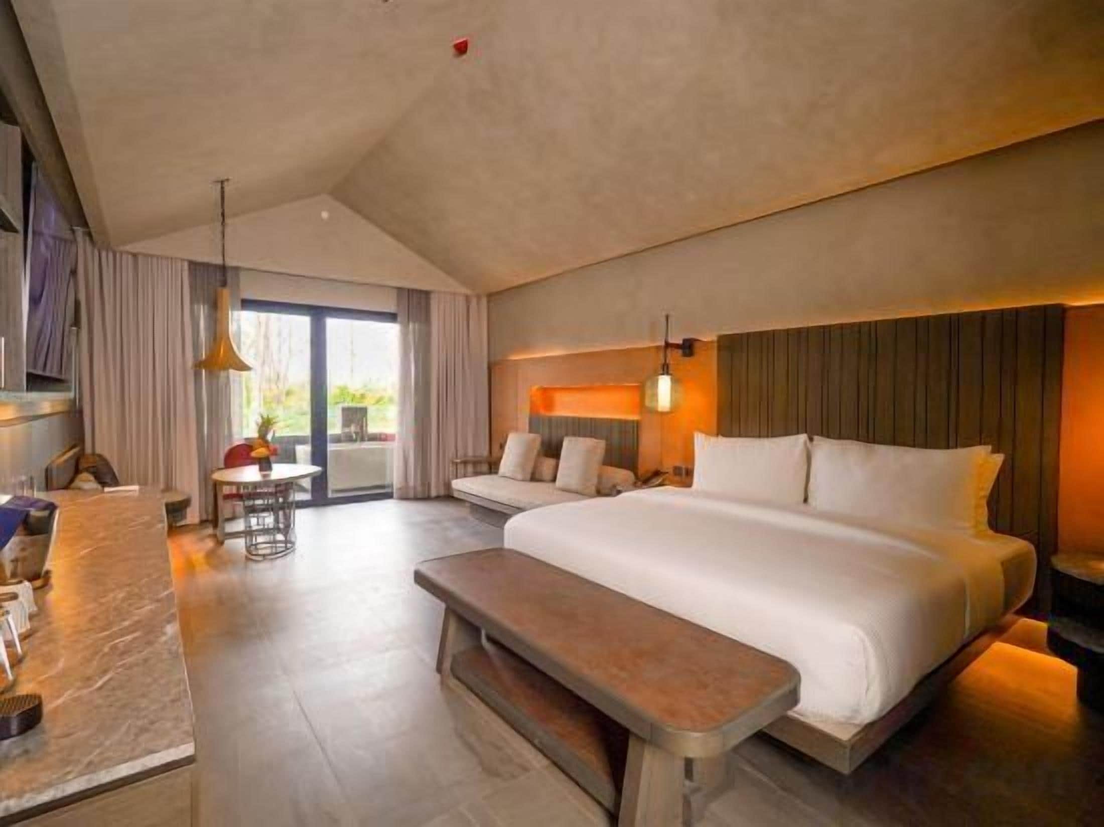 A hotel room with a large bed, a bench, a dining area, and a view of the outdoors.