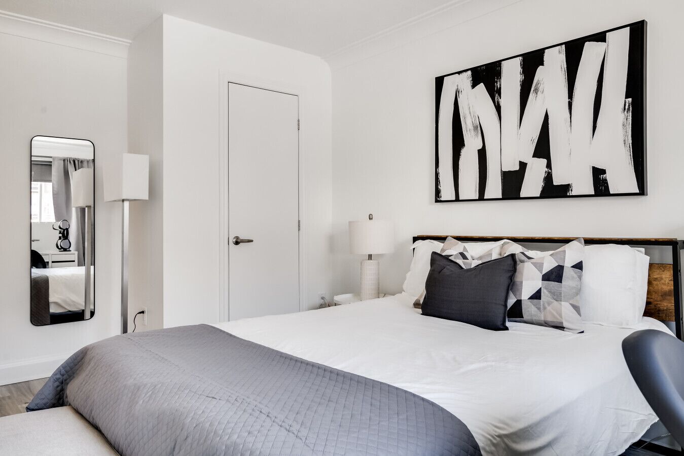 Ein Hotelzimmer mit Bett, Nachttisch, Lampe, Spiegel und einem großen, abstrakten Gemälde an der Wand.