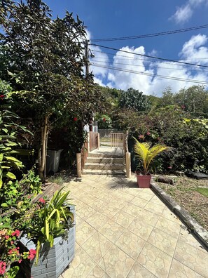 A paved pathway leading to a gated entrance, surrounded by lush greenery and flowering plants.