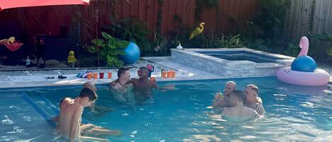 Piscina com pessoas aproveitando a água, cercada por uma cerca de madeira, um guarda-sol vermelho e diversos objetos à beira da piscina.