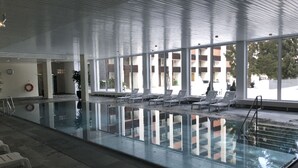 Ein Innenpool mit großen Glaswänden, Liegestühlen und Blick auf ein Gebäude.
