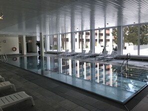 An indoor swimming pool with clear glass walls, sun loungers, and a view of a building outside. - Cristallina I, (Laax Uletsch), 37021 by Interhome (Laax)