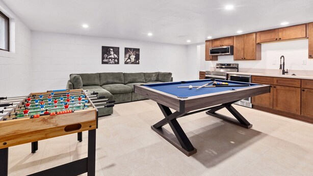 A modern living room with a foosball table, a pool table, a sofa, and a kitchen area with wooden cabinets and appliances. - Luxury 5 BR Retreat. Sleeps 10+. Near U of A. (Tucson)