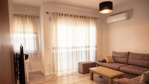 A modern living room with a sofa, ottoman, and a television. - Perla Apartment 02 in Plazhi San Pietro (Ishëm)