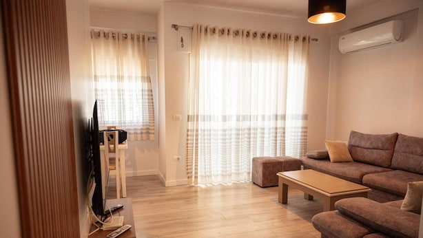 A living room with a sofa, ottoman, and a TV stand. - Perla Apartment 02 in Plazhi San Pietro (Ishëm)