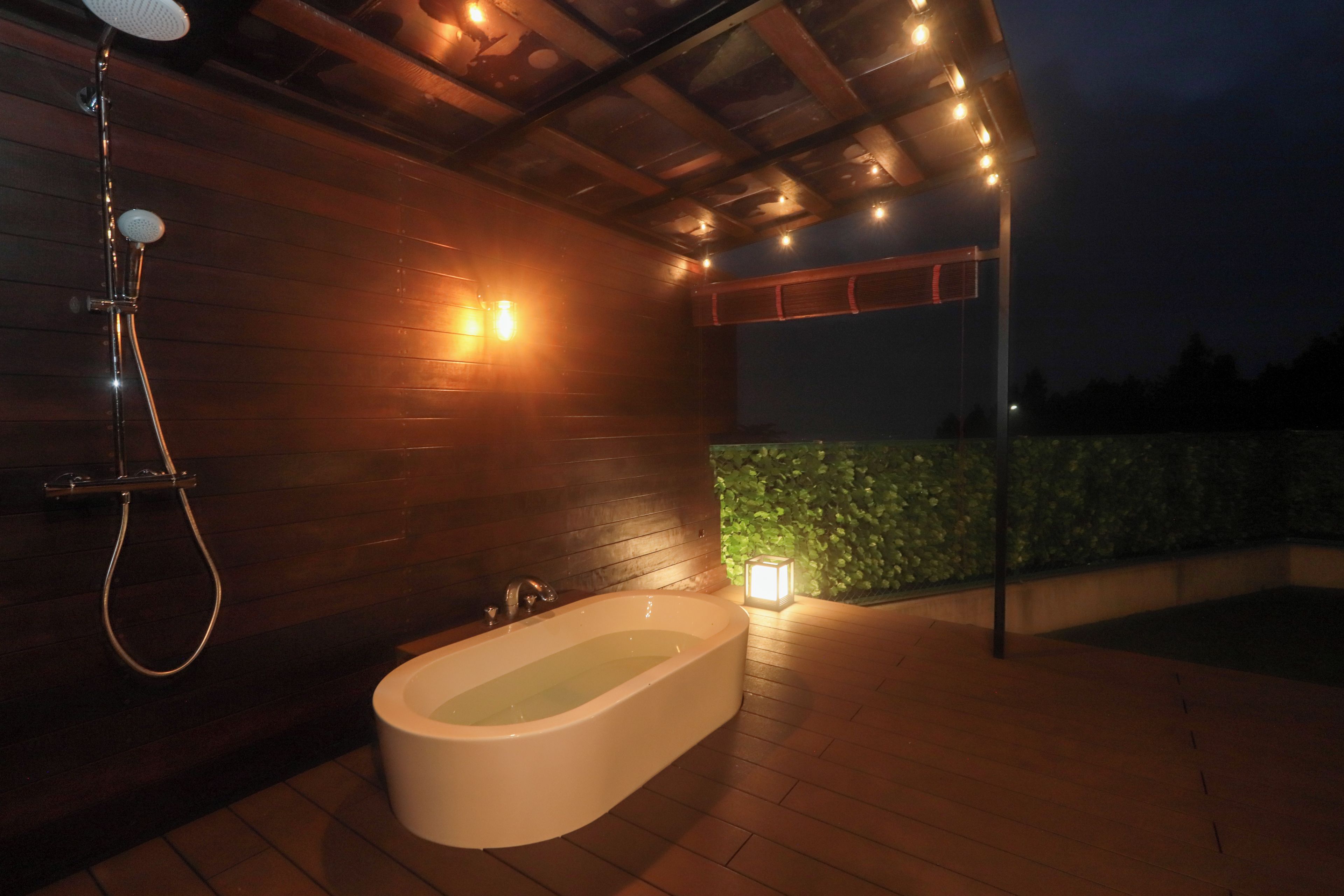 A modern outdoor bathroom with a freestanding bathtub, a showerhead, and a wall-mounted light fixture.