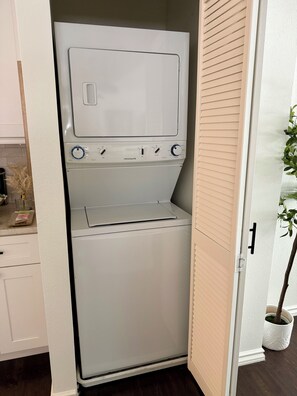 A compact, white, wall-mounted washer-dryer unit with control knobs and a front-loading door, situated in a narrow space between a white cabinet and a wooden door. - One Block From Olde Town! Hot Tub, Fire Pit, Putting Green, Arcade & Game Room (Arvada)