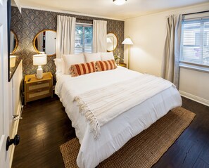 A neatly arranged bedroom with a large bed, bedside lamps, a mirror, and a window with curtains. - One Block From Olde Town! Hot Tub, Fire Pit, Putting Green, Arcade & Game Room (Arvada)