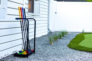 A golf cart with golf clubs and balls parked on a gravel surface next to a white building. - One Block From Olde Town! Hot Tub, Fire Pit, Putting Green, Arcade & Game Room (Arvada)