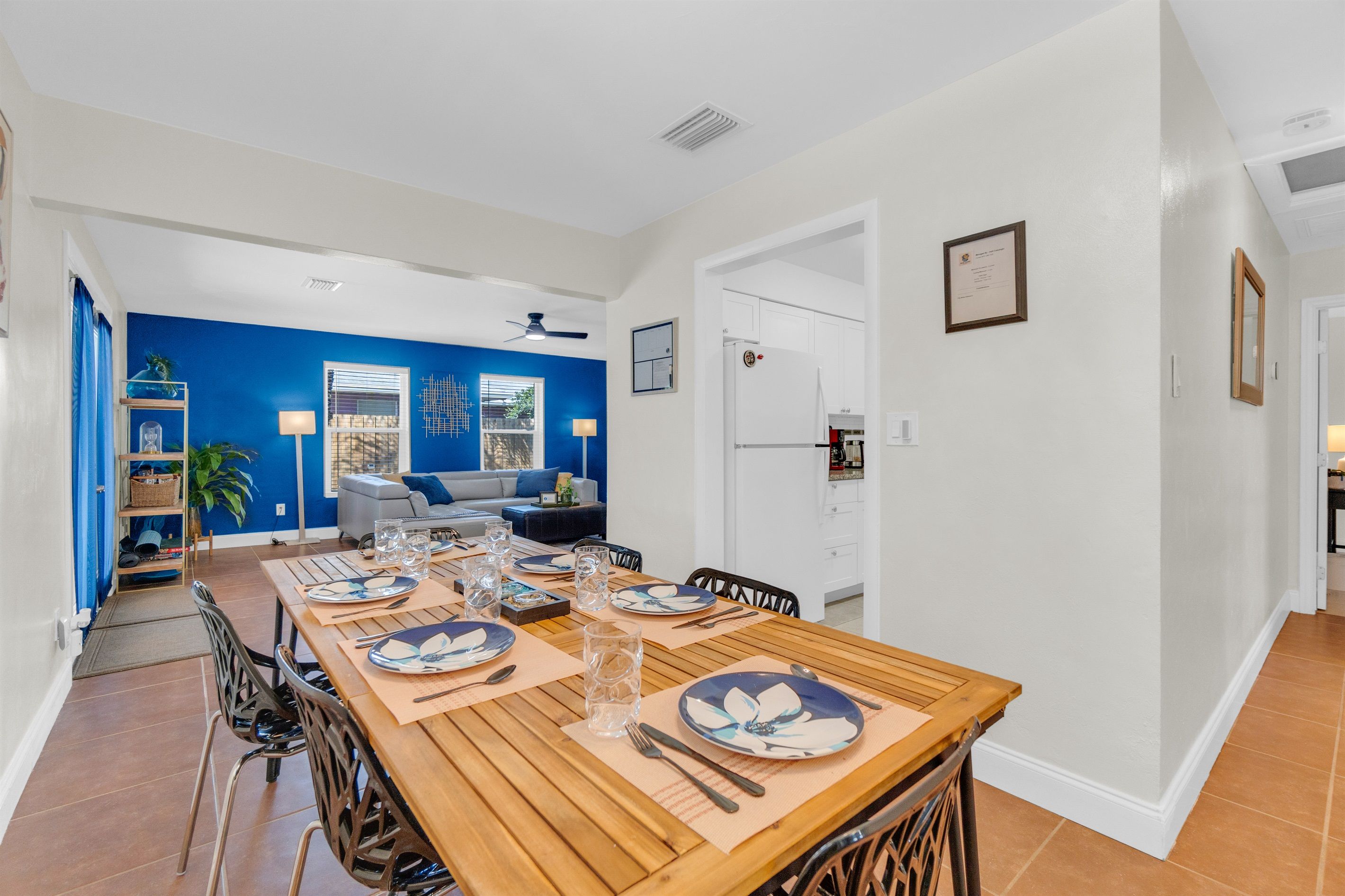 A dining area with a long wooden table set for four, a kitchen in the background, and a blue accent wall.