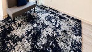 Un salon avec un canapé, un téléviseur diffusant des images de la ville, un tapis à motifs et une fenêtre avec des stores.