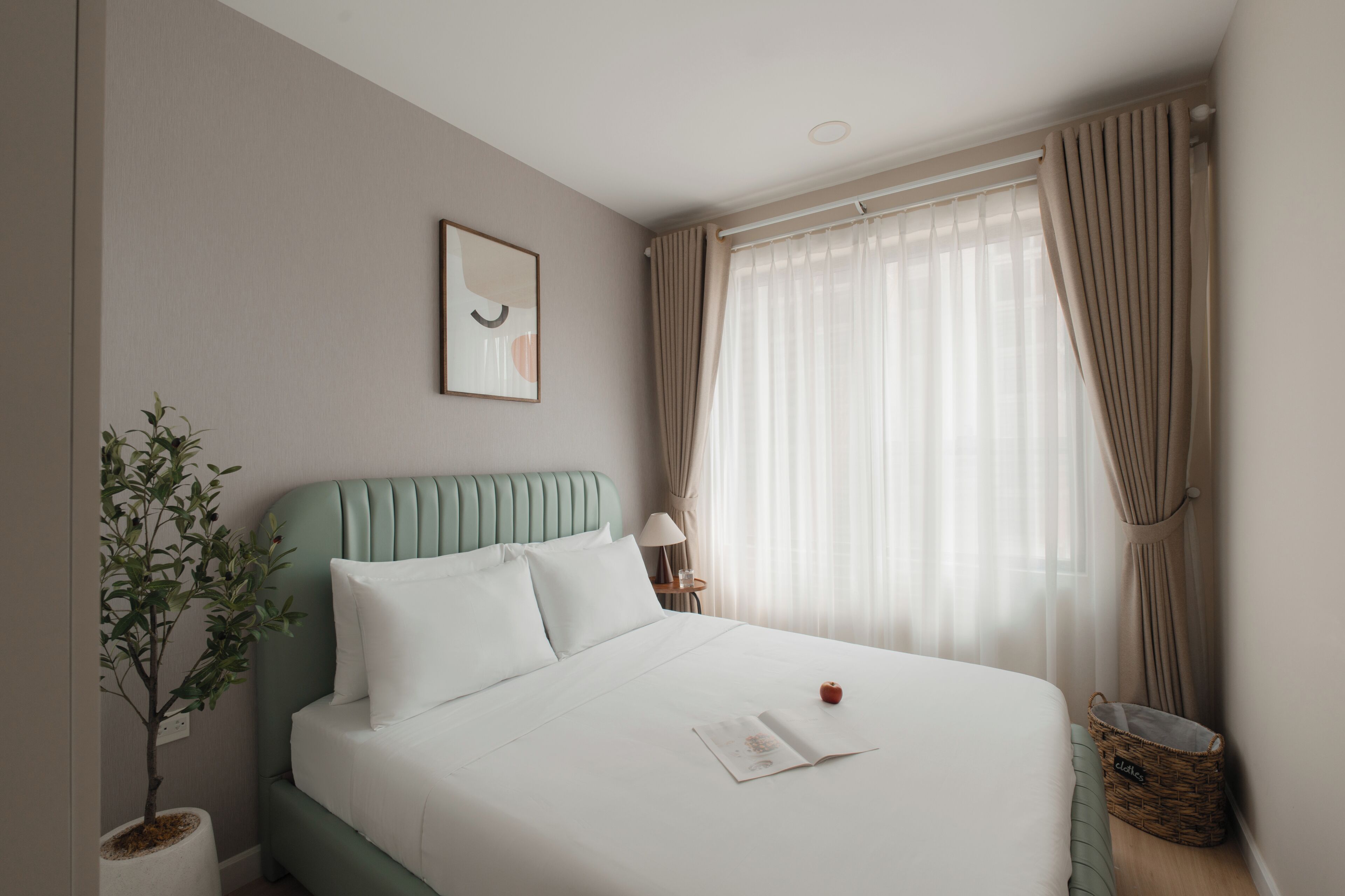 Una cama bien hecha con una almohada, una lámpara de noche, un cuadro enmarcado en la pared, una planta en maceta y una ventana con cortinas.