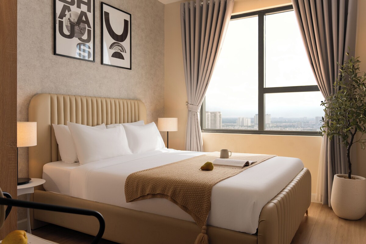 A hotel room with a large bed, a bedside table, a lamp, a window with curtains, and a potted plant.
