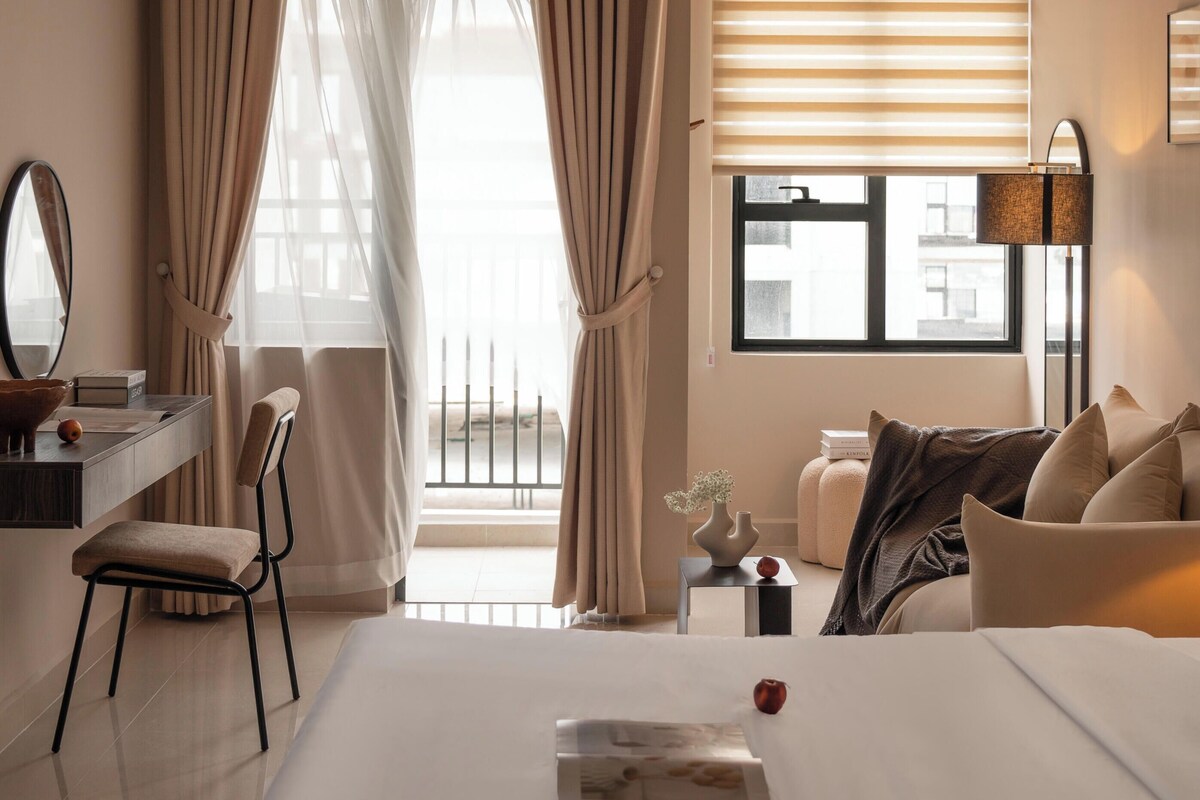 A hotel room with a bed, bedside table, chair, and a window with blinds.