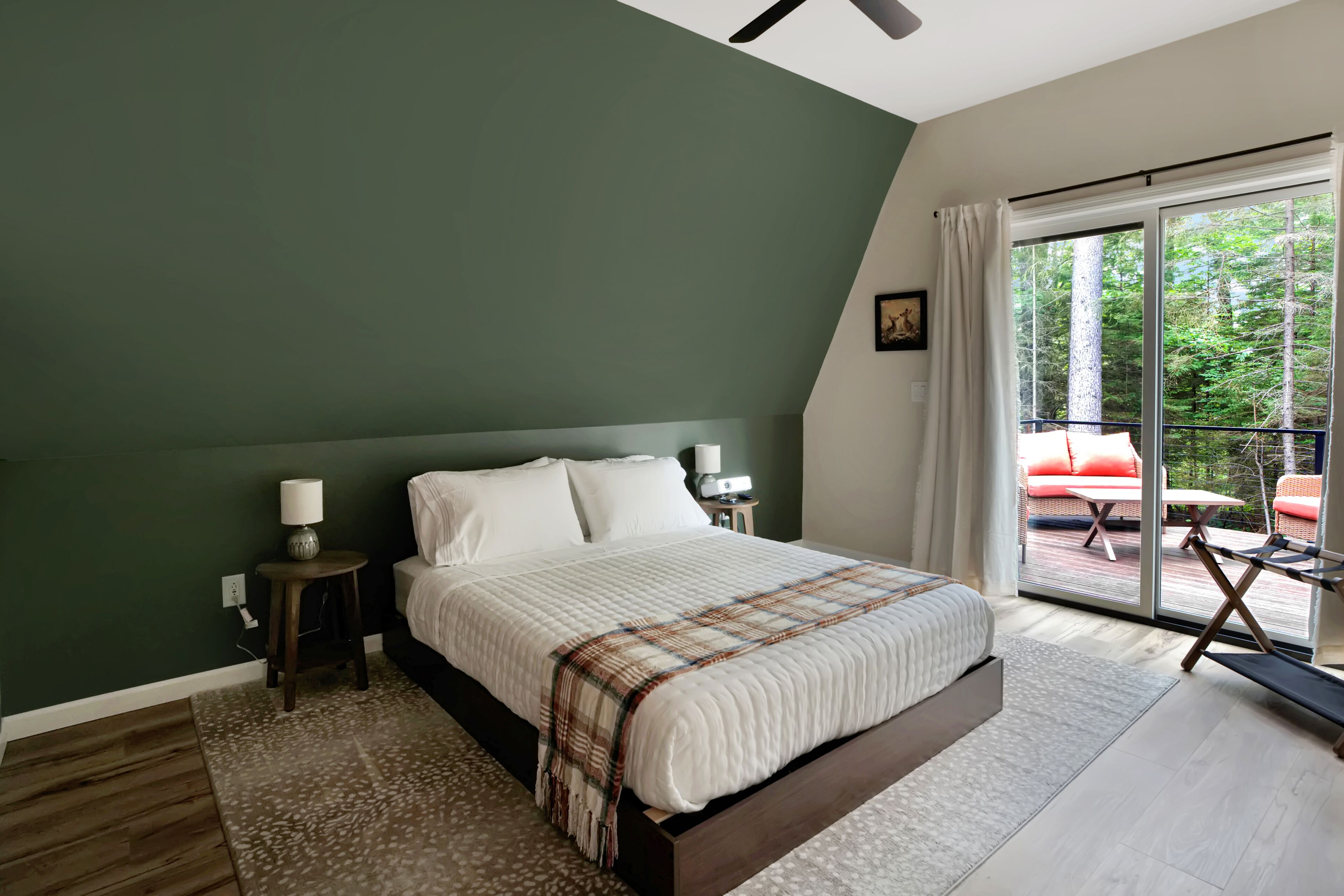 Un dormitorio con una cama grande, una mesita de noche, una lámpara, una ventana con vistas a árboles y un balcón con mobiliario exterior.