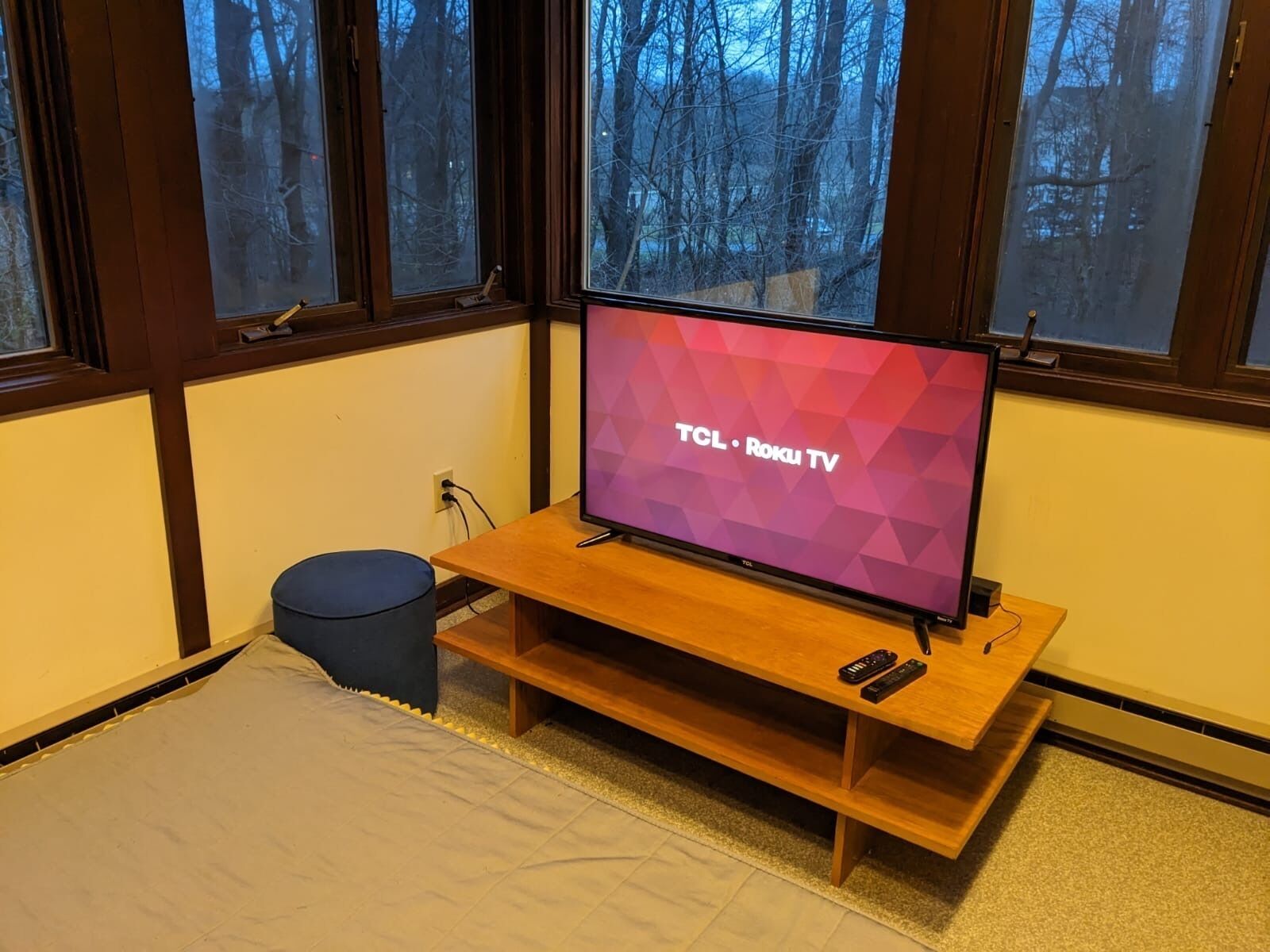 A room with a television on a wooden stand, a bed, a small blue stool, and a window with a view of trees.