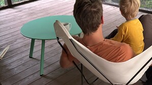 Ein Zimmer mit großem Fenster und Blick auf einen Wald. Ein grüner Tisch und zwei Stühle sind vorhanden.