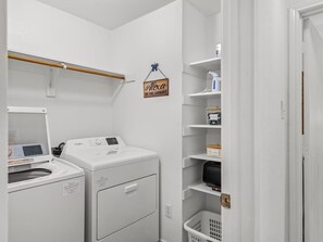 A laundry room with two front-loading washers and dryers, a drying rack, and shelves with storage boxes and a hanging sign. - Serene 3-bedroom house with AC in charming McKinney (McKinney)