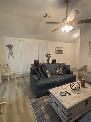A living room with a dark grey sofa, a wooden coffee table, a ceiling fan, and a window with white curtains. - Serene 3-bedroom house with AC in charming McKinney (McKinney)