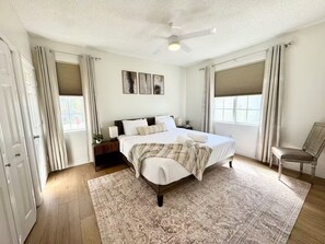 Ein Schlafzimmer mit Bett, Nachttischen, einem Sessel, einem Deckenventilator und einem Fenster mit Vorhängen.