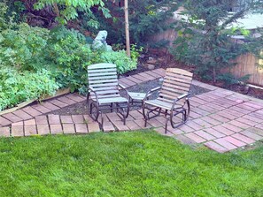 Eine Terrasse mit zwei Metallstühlen und einem Tisch, umgeben von einer Ziegelkante und üppiger Bepflanzung.