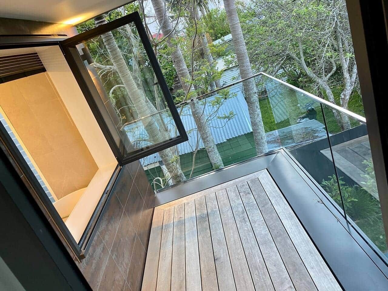 A modern balcony with a wooden floor, glass railings, and a view of trees and a building.