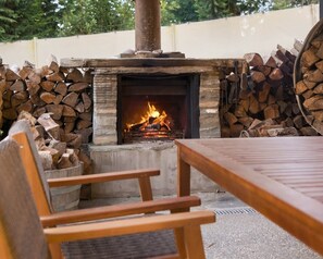 Una chimenea exterior con fuego, rodeada de leña apilada, y una mesa de madera y sillas al frente.