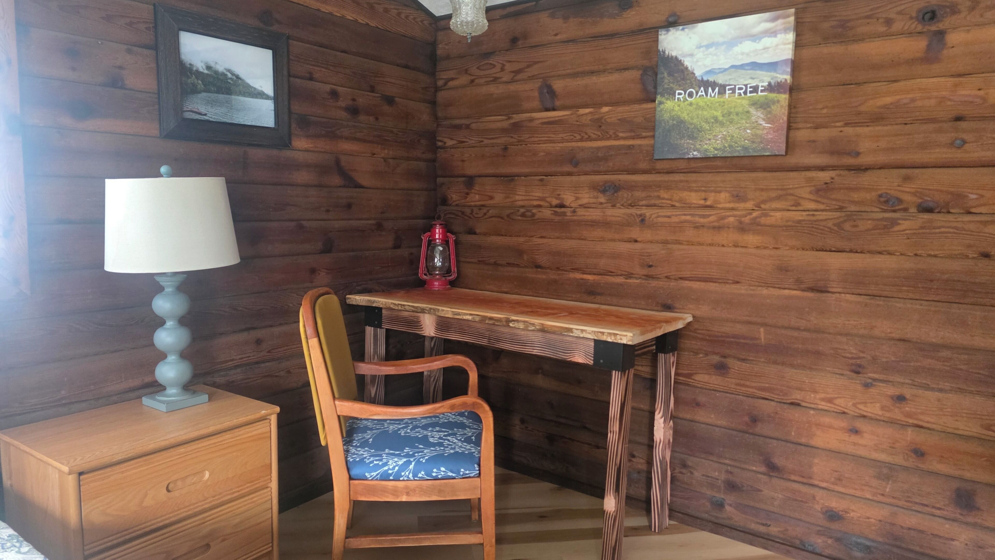 A wooden cabin interior with a desk, chair, lamp, and two framed pictures.